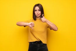 Young,woman,over,yellow,wall,making,good Bad,sign.,undecided,between