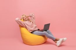 Relaxed Freelancer Lady Leaning Back In Beanbag Chair, Resting While Working Online On Laptop Over Pink Background