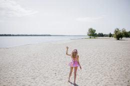 Back View To Young Blonde Girl Look Like A Barbie Doll In Pink Tones Waving Her Hand On Beach.