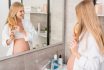 Happy Pregnant Woman In Bathrobe Brushing Her Hair And Looking At Mirror In Bathroom