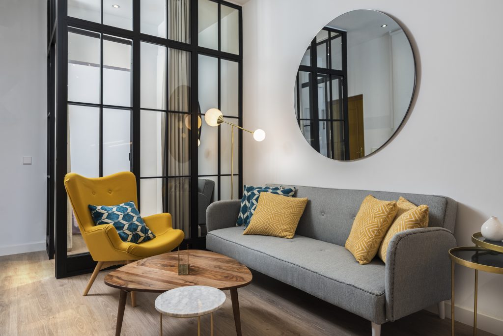 Colorful And Cozy Living Room With A Designer Armchair And Sofa Along With A Round Decorative Mirror And Glass Wall.