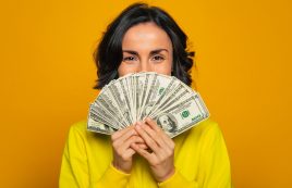 Here Is My Salary! Close Up Photo Of A Young Girl In A Yellow Hoodie, Smiling With Her Eyes, Hiding Her Face Behind Big Amount Of Money In Her Hands.