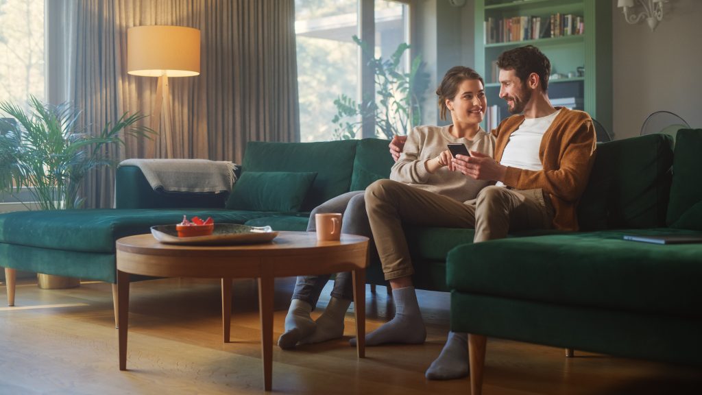 Couple Use Smartphone Device, While Sitting On A Couch In The Cozy Apartment. Boyfriend And Girlfriend Talk, Do E Shopping On Internet, Watching Funny Videos, Use Social Media, Streaming Service.