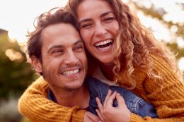 Portrait Of Happy Loving Couple With Man Giving Woman Piggyback As They Hug In Autumn Park Together