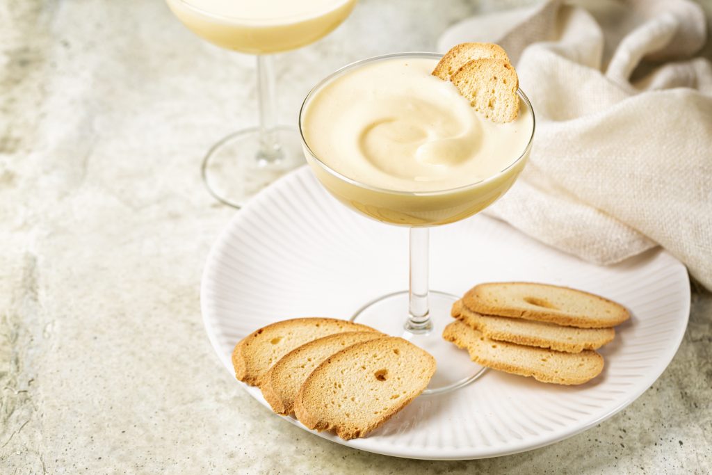 Zabaione Or Sabayon, Or Advocaat In A Champagne Coupe, Alcoholic Creamy Drink Or Dessert. Cream Made With Egg Yolks, Sugar, And A Sweet Wine Marsala Or Liqueur. Baicoli Biscuits. White Backhground, Copy Space.