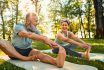 Flexible Exercises For Body. Sporty Man And Woman With Grey Hair Stretching On Yoga Mats With Hands To One Leg During Outdoors Workout. Happy Married Couple With Bare Feet Warming Up Together At Park.