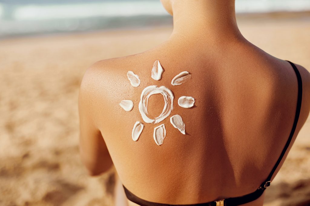 Skin Care. Sun Protection. Woman Apply Sun Cream. Woman With Suntan Lotion On Beach In Form Of The Sun. Portrait Of Female With Drawn Sun On A Shoulder. Suncream. Girl Holding Moisturizing Sunblock.