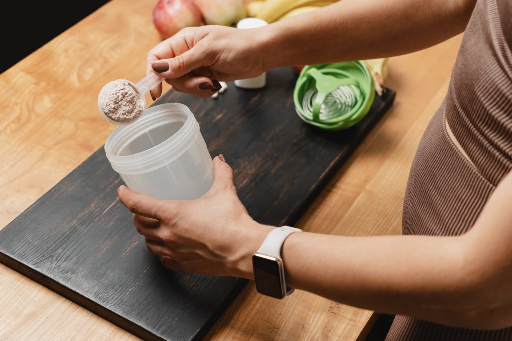 Athletic Woman In Sportswear With Measuring Spoon In Her Hand Puts Portion Of Whey Protein Powder Into A Shaker On Wooden Table With Amino Acid White Capsules, Bananas And Apple, Making Protein Drink