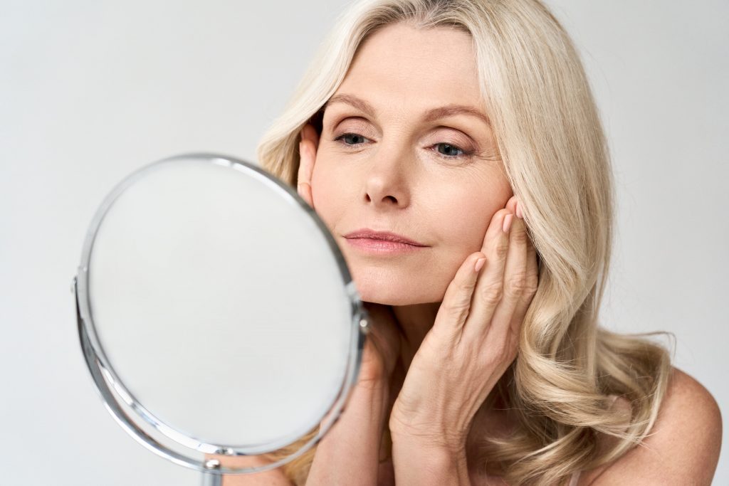 Closeup Portrait Of Gorgeous Happy Middle Age Woman Looking At Mirror Touching Her Skin Enjoying Treatment For Dry Skin. Advertising Of Antiaging Beauty Skin Care Products.