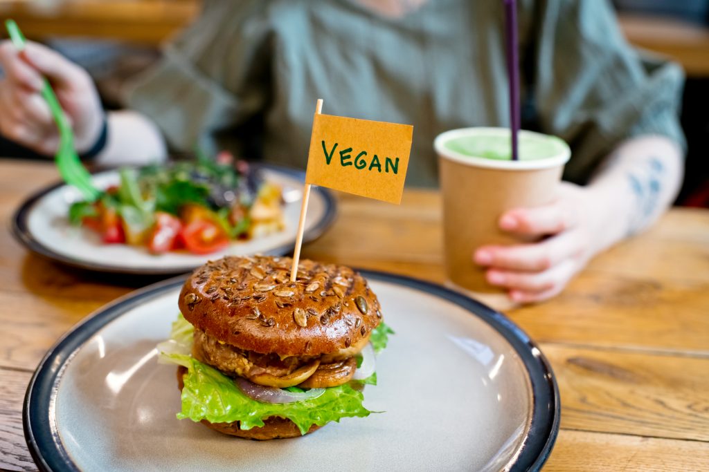 Woman Eating Veggie Salad And Burger With "vegan" Word On Small Flag In A Veggie Restaurant Or Food Court