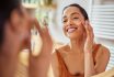 Smiling Mixed Race Young Woman Applying Moisturizer On Her Face In Bathroom