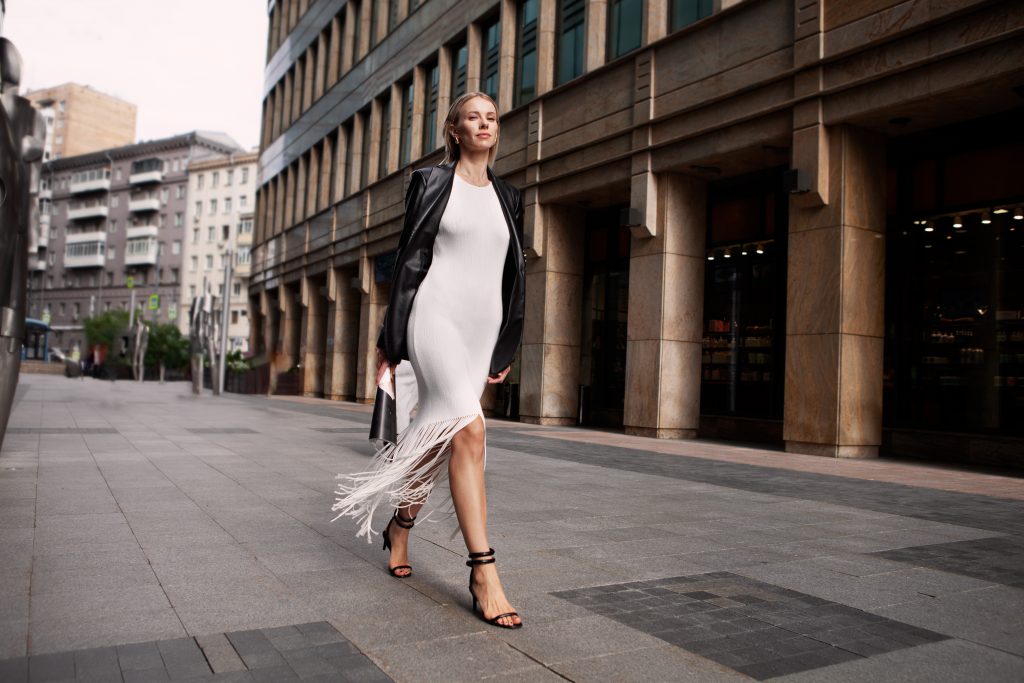 Full Length Fashionable Blonde Woman Walking City Street In Chic Attire With Building In Background. Young Female Model Wears White Fringed Dress, Black Leather Jacket And Sandals, Looks At Camera