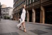 Full Length Fashionable Blonde Woman Walking City Street In Chic Attire With Building In Background. Young Female Model Wears White Fringed Dress, Black Leather Jacket And Sandals, Looks At Camera