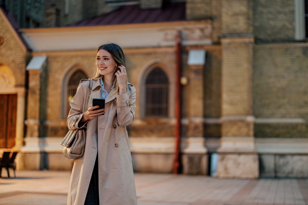 Stylish Female Walking In The City And Holding A Mobile Phone In Her Hands