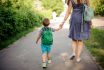 Back View Of Mother Walking Down The Street With A Little Son With A Backpack On Sunny Day