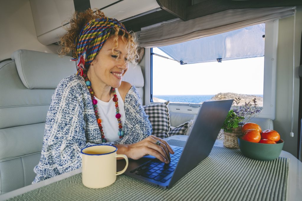 Modern Young Woman Work On Laptop Computer Inside A Camper Van Rv With Ocean Beautiful Ocean View Outside. Concept Of Online Freedom Job And Smart Working People. Digital Nomad Using Connection