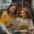 Teacher Helping A Young Student With Her Homework In The Library After School. Two Females Are Reading A Book Together In The Bookstore. They Are Doing Research For A Project