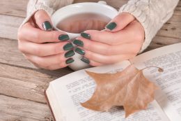 Woman Hands In Sweater With Olive Color Manicure Holds Cup Of Tea And Open Book