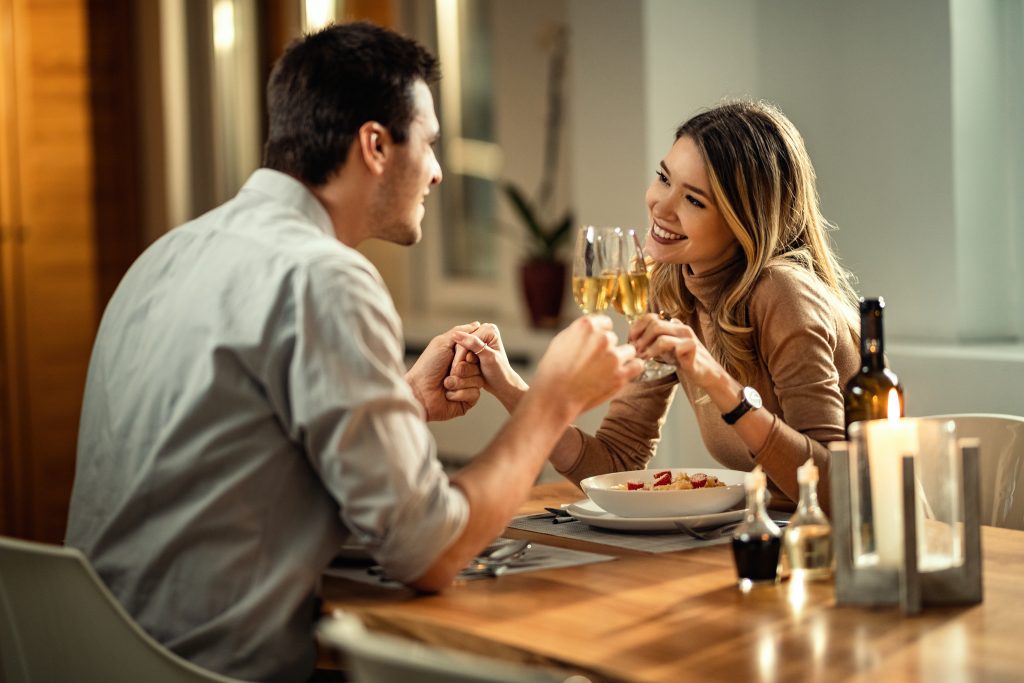 Happy Couple Toasting With Champagne During Dinner At Dining Table.