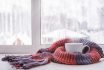 A Cup Of Hot Tea Warmed In The Knitted Scarf Is On The Windowsill Against The Winter Window