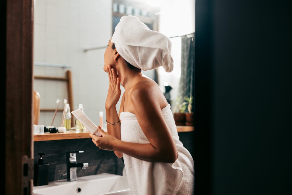 A Beautiful Woman Wrapped In A Towel Looking Herself In The Mirror While Applying Some Creme On Her Face After Taking A Bath