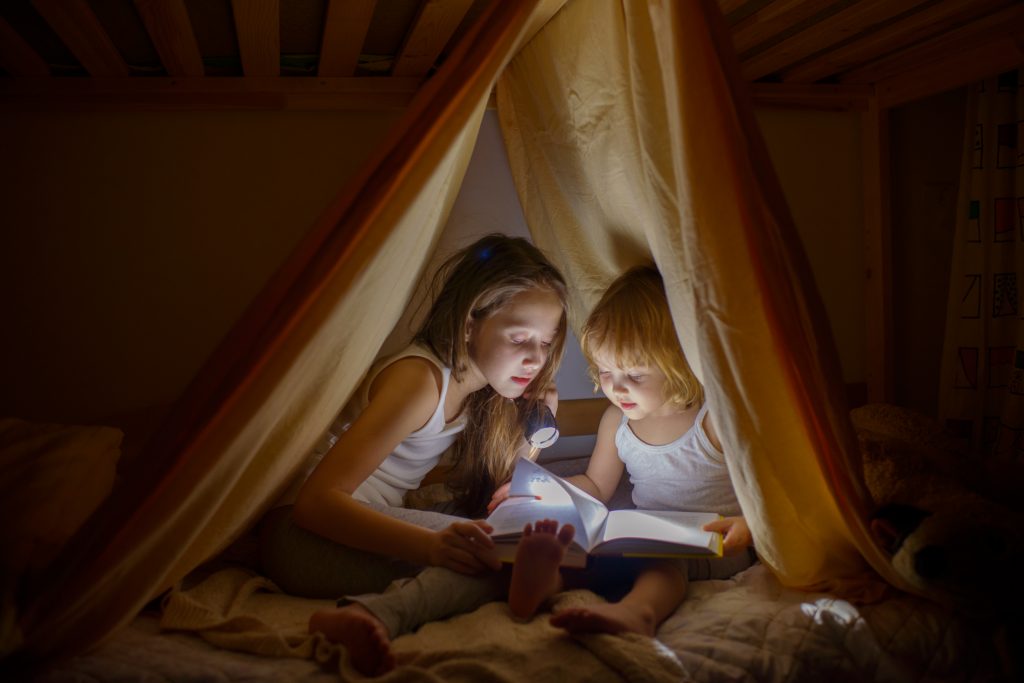 Two Girls Read Fairy Tales At Dark Night Under A Blanket By The Light Of A Small Lamp