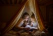 Two Girls Read Fairy Tales At Dark Night Under A Blanket By The Light Of A Small Lamp