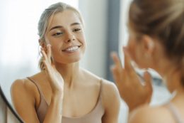 Beautiful Woman Applying Cream On Face, Standing In Bathroom At Home. Skin Care Concept, Hydration