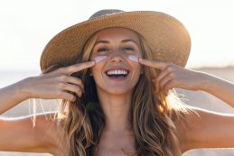 Beautiful Smiling Woman Applying Sunscreen On Her Face While Looking At Camera At The Beach.