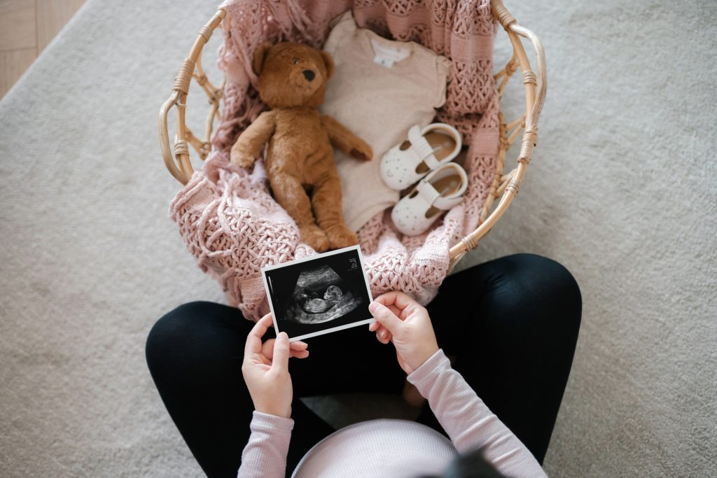 Overhead View Of Asian Pregnant Woman Holding An Ultrasound Scan Photo Of Her Baby, With A Moses Basket Filling With Baby Clothing, Baby Shoes And Soft Toy Teddy Bear In Front Of Her. Mother To Be. Expecting A New Life. Preparing For The New Born Concept