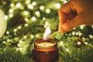 Hand Lights A Candle With A Match Against The Background Of Green Christmas Tree Branches And Lights.