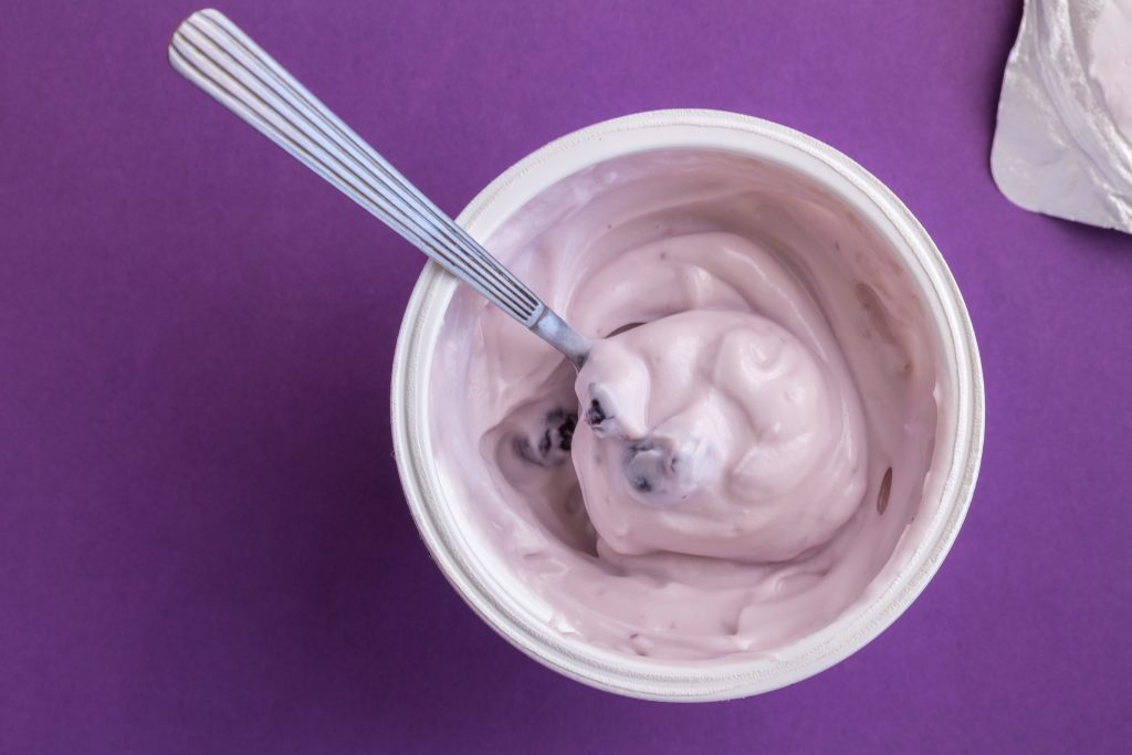 Yogurt Cup With Blue Berry Yoghurt, Spoon And Foil Lid Isolated On Purple Background