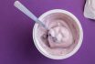 Yogurt Cup With Blue Berry Yoghurt, Spoon And Foil Lid Isolated On Purple Background