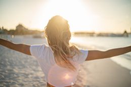 Cheerful Young Woman Embracing Nature At Sunset; Female Standing On Beach Arms Outstretched